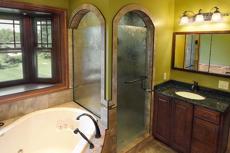 New bathroom with glass shower doors, bathtub and dark countertop and wooden cabinets.