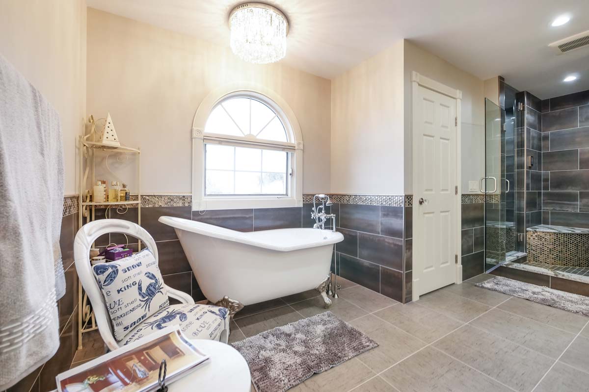 Big bathroom with a white bathtub and a glass door shower with dark tiled backsplash.