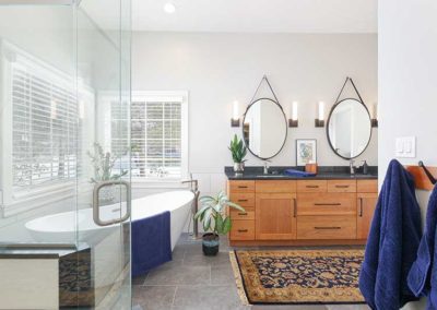 Master bathroom with a big white bathtub on the left next to windows, wooden cabinets with dark granite countertops on the right and a carpet on the tiled floor in front of the sink.